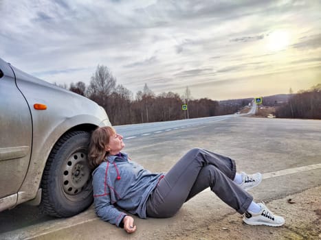 Girl Resting on Road Trip at Dusk by Her Car. A traveler takes a break, leaning on her parked car on an open road, with the sky hinting at dusk