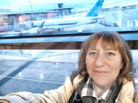 A middle-aged woman takes selfie at the window at the airport and planes in the background. Portrait of a girl on a trip