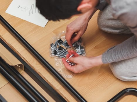 One Caucasian young man in gray clothes sits on his knees and collects screws with dowels while looking into a paper instruction for assembling furniture during the day in the room, top side close-up view with depth of field. Furniture assembly concept, assembly services.