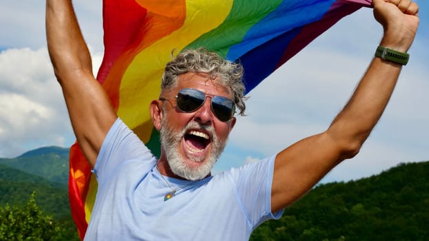 Portrait of a gray-haired elderly Caucasian man with a beard and sunglasses holding a rainbow LGBTQIA flag against a sky background, shouts in protest, Celebrates Pride Month Coming Out Day