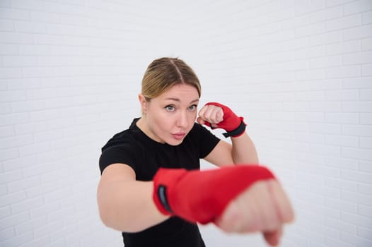 Confident waist up shot of a strong young blonde woman punching forward a camera with boxing gloves, practicing martial art, box, kickbox isolated on white background. Sport. Cardio workout. Endurance