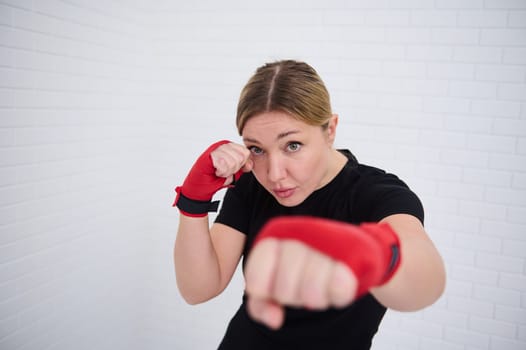 Determined female fighter with red tapes on her fists, punching forward looking at camera, isolated over white wall background. European young woman boxer 35-40 s practicing sport, boxing training