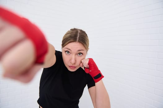 Determined female fighter with red tapes on her fists, punching forward looking at camera, isolated over white wall background. Young woman boxer 40 s practicing boxing training. Martial art combat