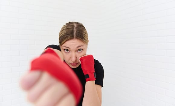 European blonde confident determined female boxer wearing boxing gloves, practicing punching forward the camera, isolated on white background. Blurred fists of hands on the foreground. Copy ad space.