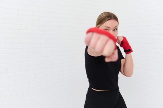 Focus on the vision of a Caucasian young female boxer fighter in red boxing gloves, punching over white isolated wall background. Caucasian confident woman exercising during box forward the camera.