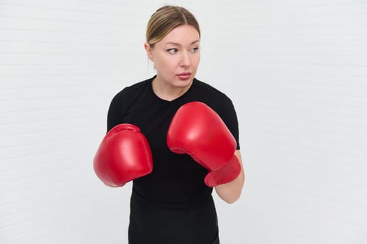 Authentic portrait of European female boxer with red boxing gloves, looking aside, isolated over white background with copy space. Pretty woman exercising during box workout indoors