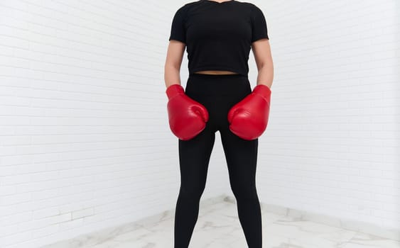 Authentic cropped portrait of a female boxer in black active wear with red boxing gloves, looking aside, isolated white background with copy space. Pretty woman exercising during box workout indoors