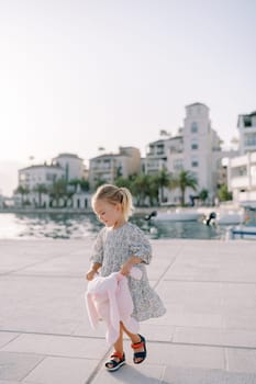 Little smiling girl walking with a pink plush rabbit on a pier by the sea. High quality photo
