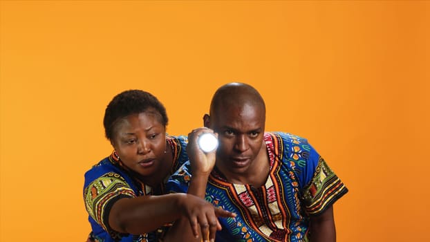 Confident man using flashlight to see what caused noise, trying to calm his wife who is hiding behind him. Terrified woman being anxious about weird sound, husband using lamp light and searching.