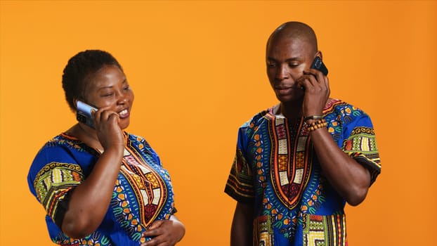 Cheerful couple talking to family on phone call, having remote conversation with smartphone over orange background. Young people in traditional clothes chatting on telephone line.