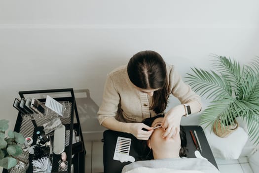 Portrait of one young beautiful Caucasian brunette girl cosmetologist gluing a paper sticker under the lower eyelid of a female client lying on a cosmetology table in the office on a spring day, close-up top view. Concept of eyelash extensions, beauty industry.