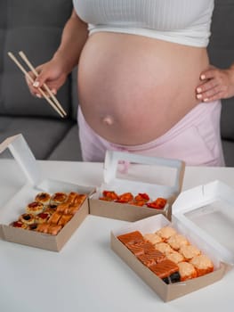 A pregnant woman holds chopsticks and stands at the table with rolls. Close-up of the belly