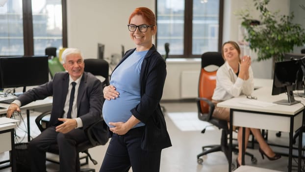 A happy pregnant woman stands in the middle of the office next to a Caucasian woman and an elderly man working at computers