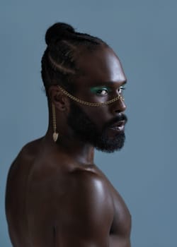 Portrait of serious African American shirtless gay man with bright makeup and gold accessory on face. Confident muscular bearded LGBTQ person looking at camera isolated on blue background.