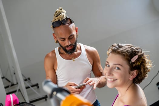 From above of gay professional make-up artist using smartphone near smiling female model looking at camera. LGBTQ person surfing on mobile phone near happy woman on backstage.