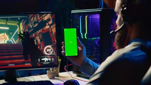 Man watching tutorial on green screen phone while playing first person shooter videogame with gun shooting laser bullets. Gamer learning to play game by looking at online guide on smartphone