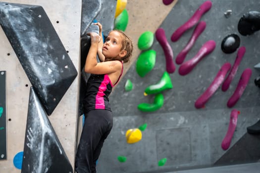 Side view of determined girl climbing artificial wall. Brave and strong female child is ascending mock wall at bouldering center, showcasing her sporty skills.