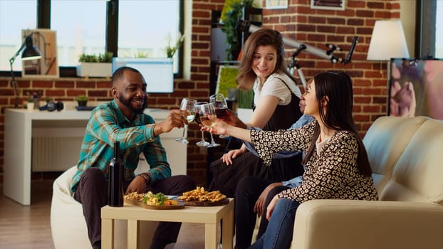 Diverse group of people celebrating their friendship together, clinking wine glasses at apartment party. Happy friends having reunion gathering, toasting alcoholic beverages