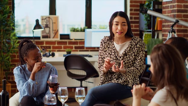 Asian woman entertaining friends at apartment party, sharing with them funny tale. Guest making multiethnic group of people laugh in living room while drinking alcoholic beverages