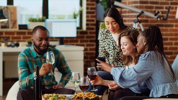 Multiracial friends at apartment party looking at social media posts made by acquaintance on cellphone, making fun of them. Amused guests in living room reacting to cringe images posted online by mate