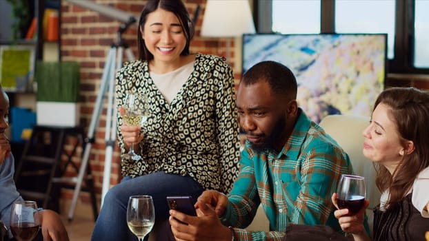 Multiracial friends at apartment party looking at funny videos on cellphone screen, sharing their impressions. Joyful guests in living room reacting to online clips on mobile phone