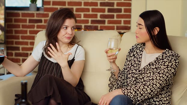 Asian woman chatting with friend on comfortable couch at apartment party, drinking wine and enjoying conversation. Joyful BFFs catching up with each other at home, consuming champagne
