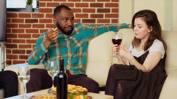 African american man flirting with woman on couch at apartment party, enjoying conversation. Guest telling pickup lines to woman at home during friends birthday, talking and drinking alcohol