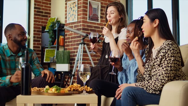 Culturally inclusive group of friends talking, drinking wine, tasting cheese and bread sticks at reunion gathering. BIPOC mates enjoying social life in brick wall living room