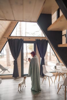 Caucasian woman wrapped in a blanket drinks coffee near the patio window hugging a Jack Russell terrier dog. Vertical photo