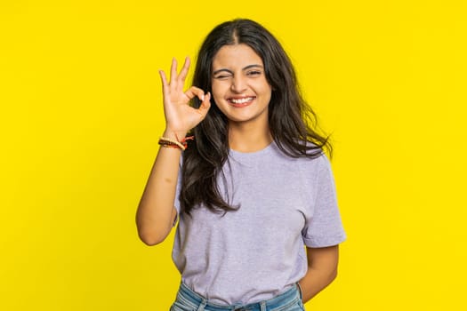 Okay. Happy Indian woman looking approvingly at camera showing ok gesture like sign positive feedback approve good choice celebrate victory wink blink eye flirting. Arabian girl on yellow background