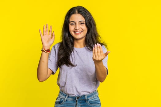 Young Indian woman smiling friendly at camera, waving hands gesturing hello greeting or goodbye welcoming with invitation hospitable expression. Arabian Hindu girl isolated on yellow studio background