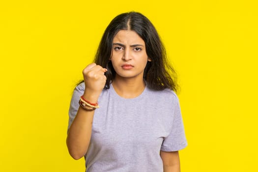 Aggressive angry Indian young woman trying to fight at camera, shaking fist, boxing with expression, punishment, threaten, bullying, abuse, mad fury. Arabian girl isolated on yellow studio background