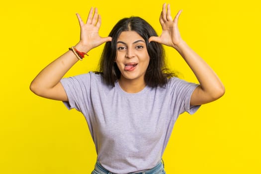 Playful Indian young woman showing tongue making faces at camera, fooling around, joking, aping with silly face, teasing, bullying, abuse. Cheerful funny Arabian girl isolated on yellow background