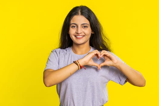 Woman in love. Smiling attractive Indian woman makes heart gesture demonstrates love sign expresses good positive feelings and sympathy. Arabian Hindu young girl isolated on yellow wall background