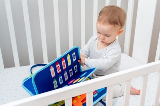 One year old baby playing with montessori busy book sitting in crib