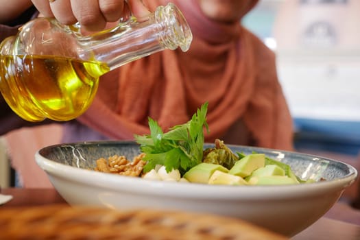 A person pouring olive oil onto a fresh, healthy salad, featuring avocados and walnuts, in a cozy restaurant setting.