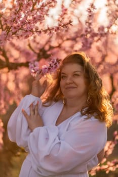 Woman blooming peach orchard. Against the backdrop of a picturesque peach orchard, a woman in a long white dress enjoys a peaceful walk in the park, surrounded by the beauty of nature