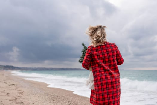 Blond woman holding Christmas tree by the sea. Christmas portrait of a happy woman walking along the beach and holding a Christmas tree in her hands. Dressed in a red coat, white dress