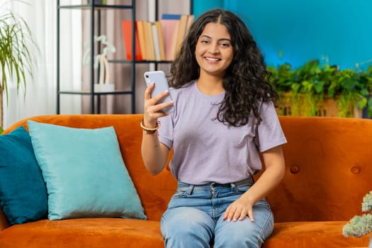 Indian woman sits on couch uses mobile phone smiles at home living room apartment. Young Arabian girl texting share messages content on smartphone social media applications online watching relax movie