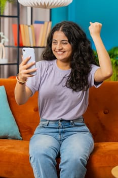 Happy woman use mobile smartphone typing browsing, celebrating success victory, winning lottery jackpot goal achievement play game good positive news, triumph. Girl at home in room on sofa. Vertical