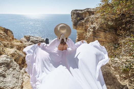 A woman in a white dress is standing on a rocky beach with her hat on. The scene is serene and peaceful, with the woman enjoying the view of the water.