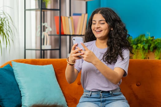 Indian woman sits on couch uses mobile phone smiles at home living room apartment. Young Arabian girl texting share messages content on smartphone social media applications online watching relax movie