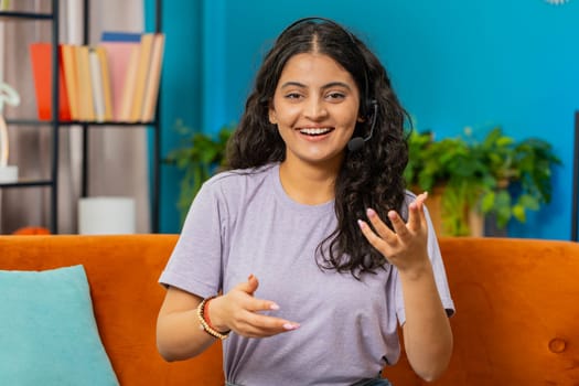 Indian woman wearing headset, freelance worker, call center or support service operator helpline. Arabian girl having talk with client or colleague communication support waving hello alone at home