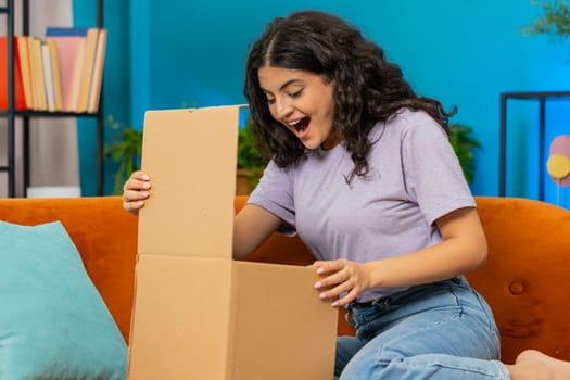 Happy Indian young woman unpacking delivery parcel sits at home. Smiling satisfied Arabian Hindu girl shopper online shop customer opening cardboard box receive purchase gift by fast postal shipping