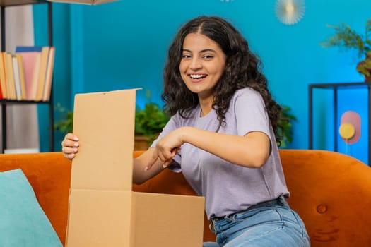 Smiling satisfied Arabian Hindu girl shopper online shop customer opening cardboard box, receive purchase gift by fast postal shipping. Happy Indian young woman unpacking delivery parcel sits at home