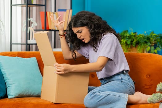 Angry dissatisfied shopper Indian woman unpacking parcel feeling upset and confused with wrong mistake delivery from an online store, bad quality broken purchase at home. Girl indoors in room on couch