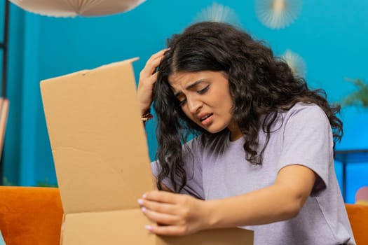 Angry dissatisfied shopper Indian woman unpacking parcel feeling upset and confused with wrong mistake delivery from an online store, bad quality broken purchase at home. Girl indoors in room on couch