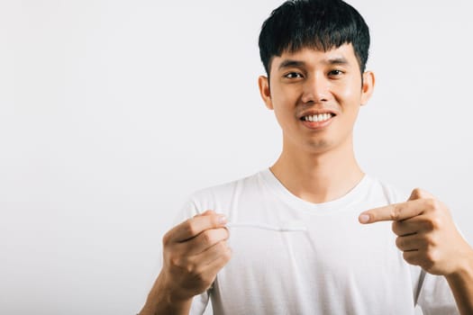 Smiling Asian teen brushes teeth in the morning, holding a toothbrush and pointing to it in a studio shot isolated on white background. Dental health concept with positive expression.