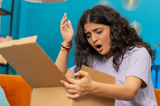 Angry dissatisfied shopper Indian woman unpacking parcel feeling upset and confused with wrong mistake delivery from an online store, bad quality broken purchase at home. Girl indoors in room on couch