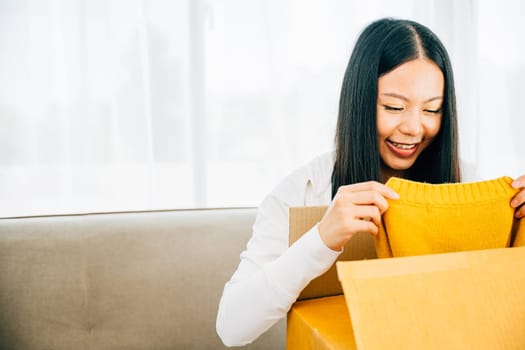 On a couch an Asian woman unpacks a delivered shirt from online shopping. Her smile signifies happiness and satisfaction with the dress. Satisfied buyer concept.